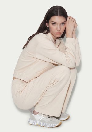 Young woman in beige tracksuit crouching, resting head on hands, wearing gray and white sneakers with gum soles.