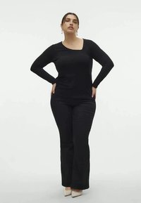 Black long-sleeve top with a square neckline, paired with fitted black pants, both made of smooth fabric. Model poses with hands on hips.