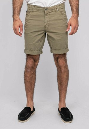 Homme portant un short marron clair retroussé, une chemise blanche et des chaussures noires slip-on, debout devant un fond clair uni.