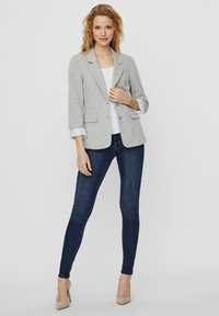 Blazer gris avec un col à revers, deux poches à l'avant et des manches retroussées. Porté sur un haut blanc et associé à un jean skinny bleu foncé et des talons.