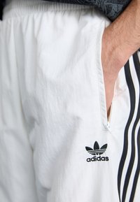 Hvite sportsshorts med svart Adidas-logo. Har sidelommer og svarte vertikale striper. Materialet virker lettvekts med lett tekstur.