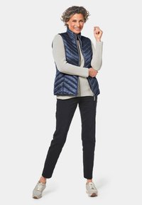 Marineblau gesteppte Weste mit hohem Kragen über einem hellbeigen Pullover, kombiniert mit dunklen Hosen und hellen Turnschuhen, mit einem Reißverschluss.