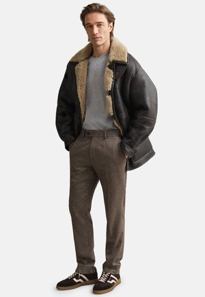 Veste en cuir noire avec col en shearling, t-shirt gris, pantalon en tweed marron et baskets marron avec des accents blancs et des lacets.