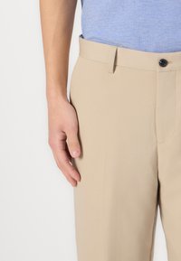 Beige nadrág sima textúrával, lapos elejével, övhurkokkal és gombos derékrésszel, kék felsővel párosítva.