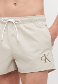 Béžové plážové šortky s elastickým pásom a šnúrkou, pričom na dolnej ľavej strane majú malé hnedé logo "ck".