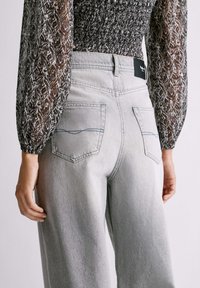 Svijetlosive traperice visokog struka s opuštenim krojem, sadrže dva stražnja džepa, diskretne detalje šivanja i crnu oznaku brenda na struku.