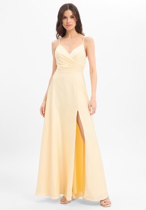 Vrouw draagt een mouwloze lichtgele maxi-jurk met V-hals, overslag lijfje, hoge split en open teensandalen, staand tegen een witte achtergrond.