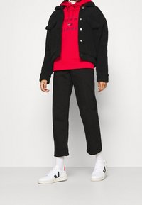 Giacca sherpa nera sopra un hoodie rosso con logo, pantaloni neri e sneakers bianche con accenti rossi e neri.