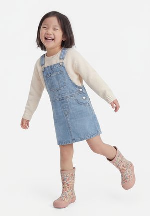 Smilende ung pige iført en cremefarvet sweater, denim overallskjole og blomstrede gummistøvler, står på ét ben med armene strakt ud for at holde balancen.