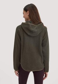 Grüner Fleece-Hoodie mit weicher Textur und Kapuze. Verfügt über einen abgerundeten Saum und subtile Nähte. Wird mit dunklen Leggings kombiniert.