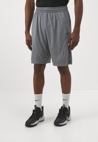 Pantalones cortos atléticos de malla gris con una cinturilla elástica, bolsillos laterales y detalles a rayas. Llevados con calcetines blancos de Nike y zapatillas deportivas negras.
