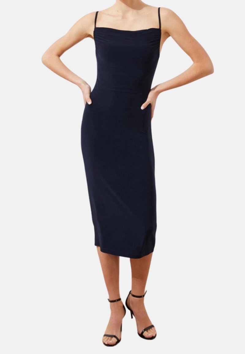 Femme portant une robe midi ajustée, sans manches, bleu marine avec de fines bretelles et des sandales noires à talons hauts, debout les mains sur les hanches.