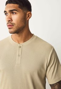 Camisa henley de canalé beige con escote redondo, tapeta de dos botones y mangas cortas, con detalles de costura visibles.