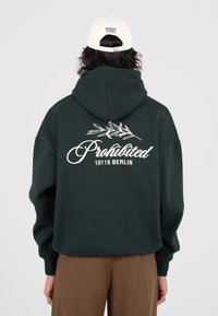 Persona che indossa una felpa verde scuro con la scritta "Prohibited 10119 Berlin" e un motivo con foglie sul retro, un cappellino beige con la scritta "Modern Yuppies" e pantaloni marroni.