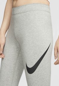 Šedé legíny Nike mají široký pas, hladkou texturu a výrazné černé logo swoosh na levém stehně.