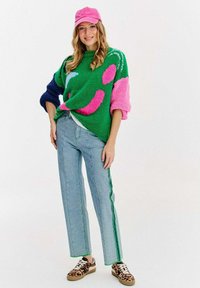 Grüner Strickpullover mit pinken und marineblauen Akzenten, der grafische Muster zeigt. Hellblaue Jeans mit grünen Seitenstreifen und Sneakers im Tierdruck.