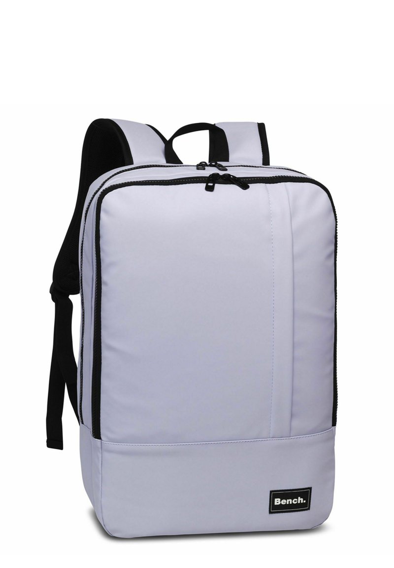 Mochila de color púrpura claro con acentos negros, forma rectangular redondeada, correas acolchadas, cierre de doble cremallera y una etiqueta de logo en el frontal.
