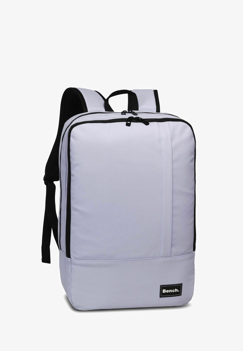 Mochila de color púrpura claro con acentos negros, forma rectangular redondeada, correas acolchadas, cierre de doble cremallera y una etiqueta de logo en el frontal.