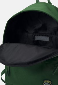 Sac à dos en tissu vert avec un intérieur noir, doté d'une fermeture éclair lisse et d'un patch logo Lacoste. Conçu pour un usage quotidien avec des bretelles réglables.
