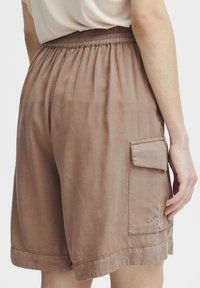 Shorts marron arrivant au genou, fabriqués en tissu léger. Dotés d'une taille élastique et d'une poche cargo sur le côté. Texture lisse, design décontracté.