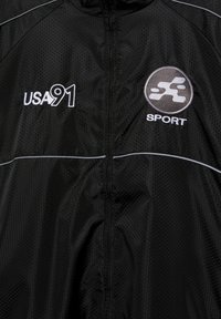Černá sportovní bunda s medovou strukturou, bílý nápis "USA 91" vlevo a kulaté logo "SPORT" na pravé hrudi.