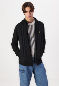 Sudadera negra con cremallera y capucha, con un pequeño logo amarillo en el pecho, usada sobre un suéter a rayas y combinada con pantalones cargo de mezclilla azules.