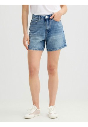 Persona que lleva pantalones cortos de mezclilla azules, zapatillas blancas y una camisa blanca, de pie con una mano en el bolsillo sobre un fondo liso.