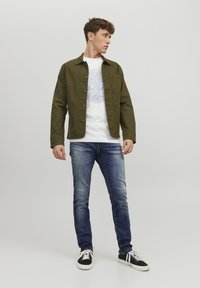 Giacca verde oliva con bottoni, t-shirt bianca con grafica blu, jeans blu e sneakers nere con lacci bianchi.