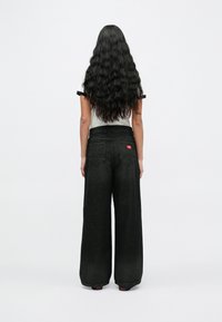 Γυναίκα με μακριά κυματιστά μαύρα μαλλιά που κοιτάζει προς τα πίσω, φορώντας λευκό κοντομάνικο πουκάμισο και ψηλόμεσα ευρεία μαύρα τζιν παντελόνια.