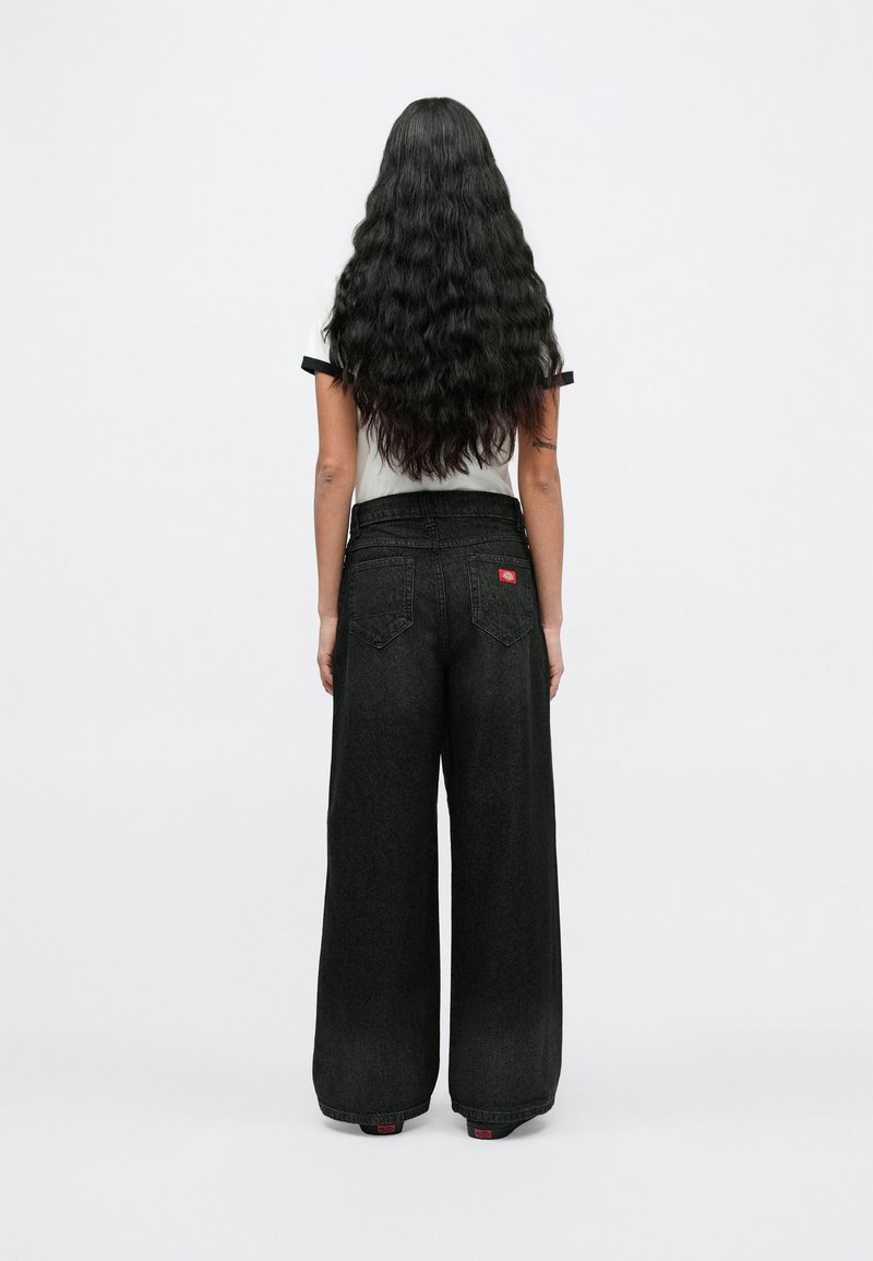 Frau mit langen, welligen schwarzen Haaren, die von hinten zu sehen ist, trägt ein weißes kurzärmeliges Hemd und hoch taillierte, weit geschnittene schwarze Jeans.