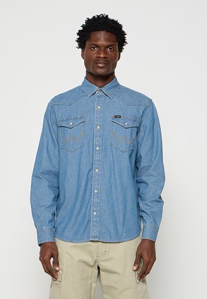 Uomo che indossa una camicia di jeans azzurro chiaro con due tasche sul petto e pantaloni cargo beige, in piedi su uno sfondo grigio uniforme.