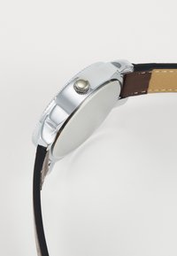 Montre en acier inoxydable argenté avec un boîtier rond, un bracelet en cuir noir et une couronne texturée ; présente un cadran lisse et vierge.