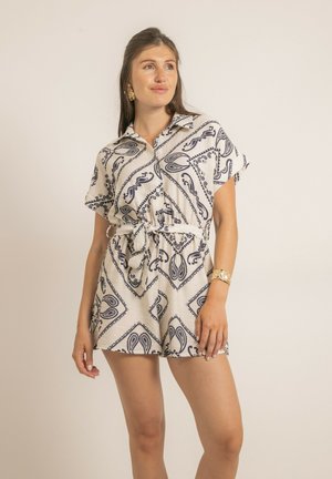 Femme aux longs cheveux bruns portant une combinaison courte à manches courtes blanche et marine à motifs, avec une ceinture nouée à la taille et des bijoux en or, debout devant un fond uni.
