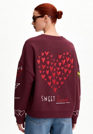 Frau mit roten Haaren im Dutt trägt eine Sonnenbrille und ein weinrotes Sweatshirt mit roten Herzmotiven und den Worten "Sweet heart" auf dem Rücken.