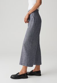 Pantalons larges en tissu texturé bleu et gris, présentant une taille haute et des lignes épurées. Associés à des mocassins noirs.
