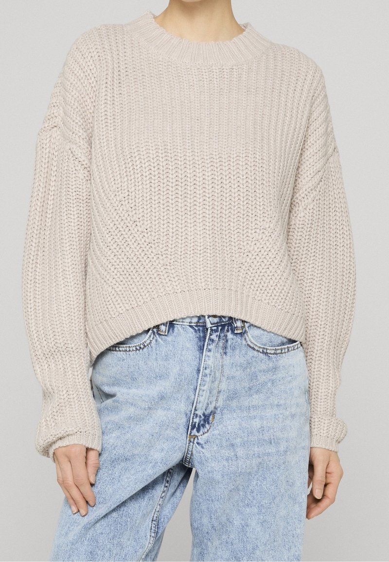 Personne portant un pull épais en maille crème à manches longues et un jean bleu clair taille haute sur un fond uni.