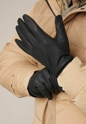 Personne portant une veste beige enfile des gants en cuir noir lisse, centrée sur un gros plan des mains et des textures des vêtements.