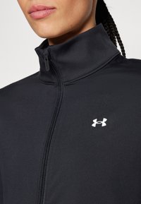 Chaqueta deportiva negra con cuello alto, cierre de cremallera y un pequeño logo blanco de Under Armour en el pecho. Material suave y ligero.