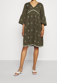 Khaki vihreä mekko V-päällä, kolmen neljänneksen liehuvilla hihoilla, brodeeratuille timanttimalleille ja aallotetulle helman. Yhdistetty valkoisten sandaalien kanssa.