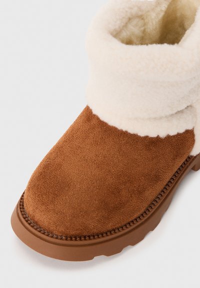 Botte cheville en daim marron avec un épais revers en fourrure beige et une semelle en caoutchouc, conçue pour la chaleur et un style décontracté.