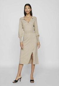 Beige gebreide jurk met een V-hals en lange pofmouwen, met een halflange split aan één kant. Gepaard met zwarte open teen hakken.
