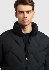 Zwarte pufferjack met hoge kraag, zichtbare rits en gewatteerde textuur. Draagt over een zwart T-shirt met ronde hals, met een modern ontwerp.