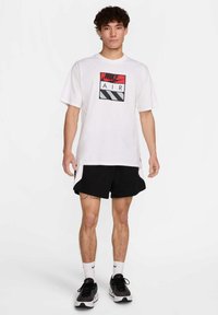 Bela kratka majica s kratkimi rokavi z rdečim in črnim grafičnim logotipom "NIKE AIR" spredaj, v kombinaciji s črnimi športnimi kratkimi hlačami in čevlji.