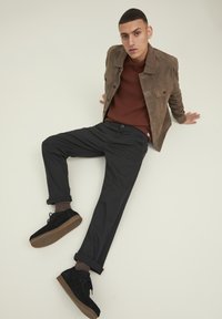 Chaqueta de ante marrón sobre un suéter de punto en color óxido, combinada con pantalones negros enrollados y zapatos de ante negros con una suela color beige, sobre un fondo neutro.