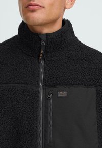 Man wearing black textured fleece jacket with high collar and zippered chest pocket labeled "Solid Denmark Since 1999."