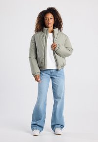 Chaqueta de color verde oliva acolchada con un alto cuello, cremallera frontal y puños elásticos, combinada con jeans de pierna ancha de color azul claro y zapatillas blancas.