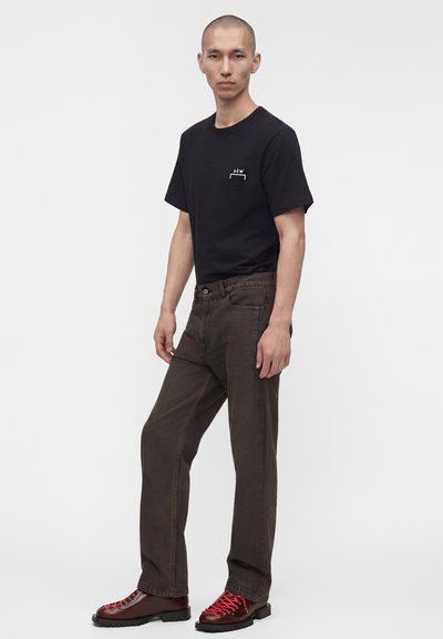 Νέος άνδρας φορώντας μαύρο t-shirt A-COLD-WALL*, σκούρα καφέ τζιν και σκουροκόκκινα μποτάκια με κόκκινα κορδόνια, στέκεται μπροστά σε λευκό, απλό φόντο.