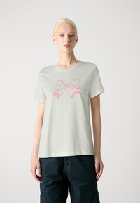T-shirt vert clair à manches courtes en tissu doux, avec un grand graphisme de nœud rose sur le devant. Encolure ronde simple.