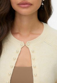 Cardigan crème en tissu doux, avec un col rond, des boutons en perles et une petite découpe au centre.