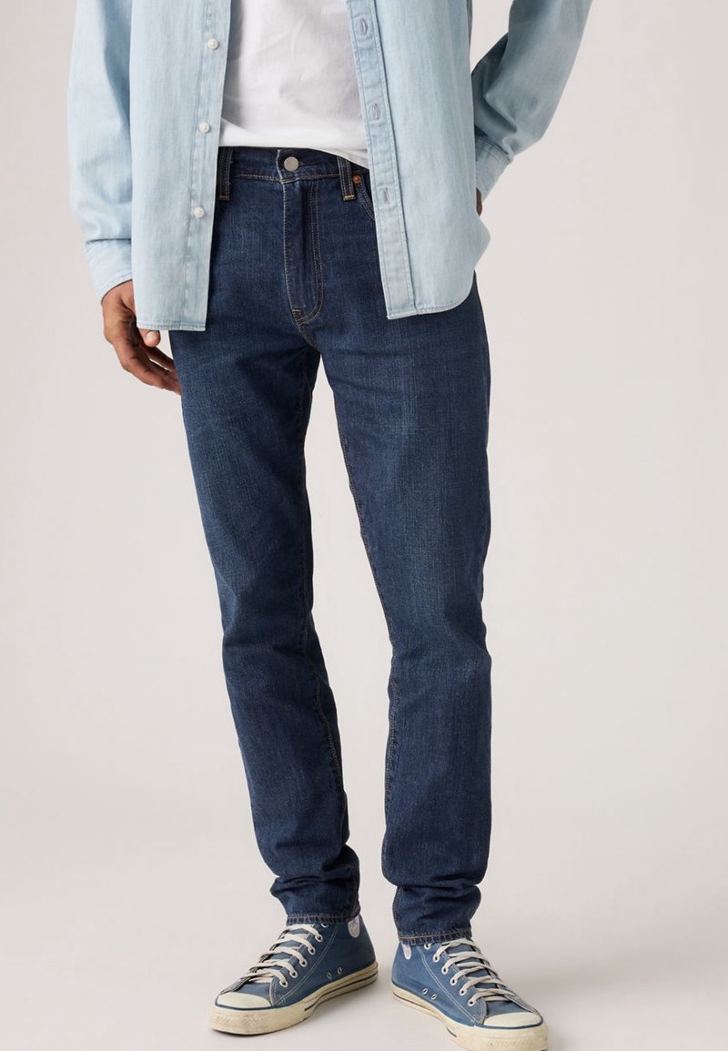 Homme portant un jean bleu foncé, une chemise en denim bleu clair, un t-shirt blanc et des baskets en toile bleues, debout sur un fond uni.